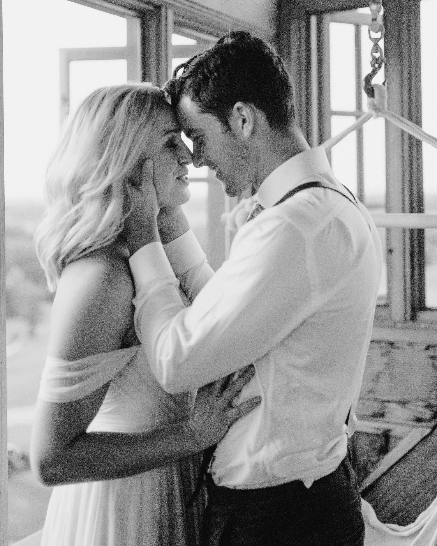 A romantic black and white photograph of a couple in an intimate moment sharing affection by a window.