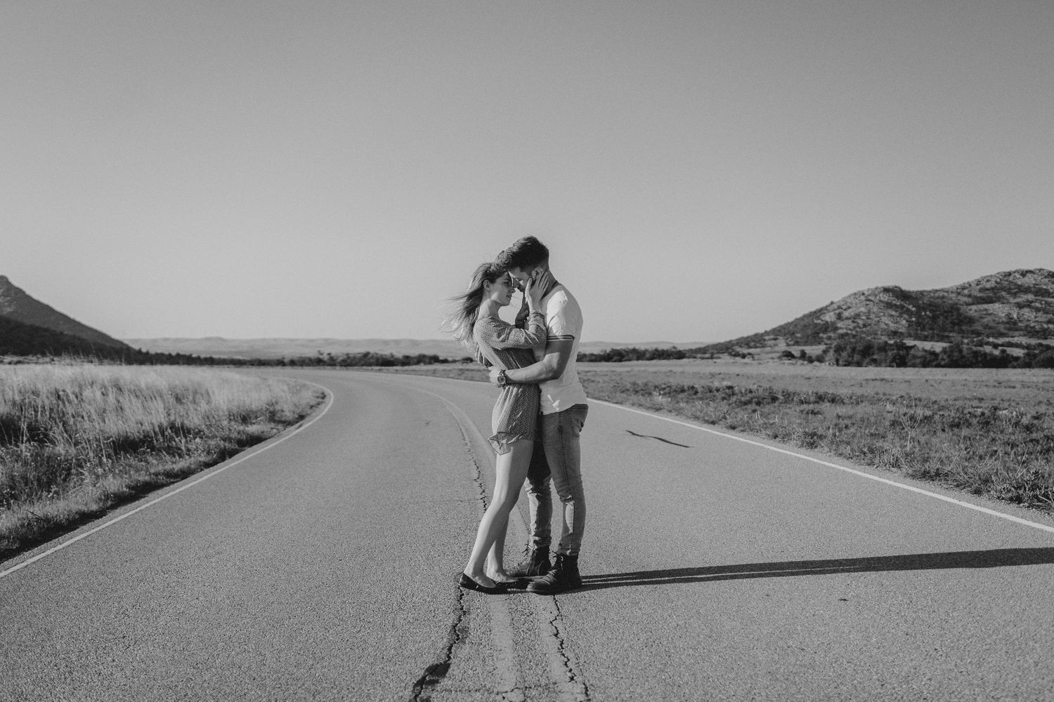A couple embraces while standing in the middle of an empty rural road with mountains in the distance in black and white.