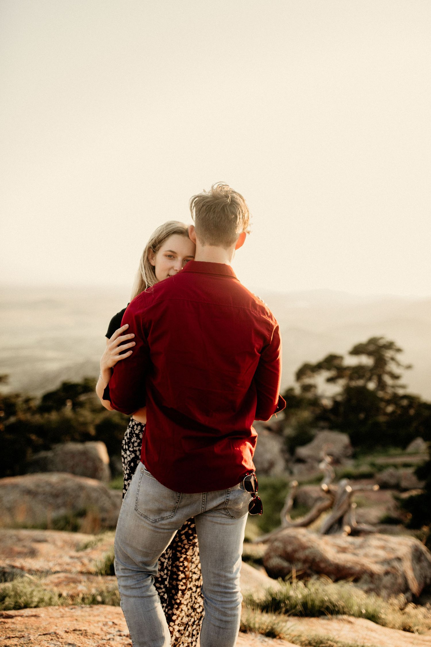 Couple embraces on rocky hillside during golden hour with scenic mountain landscape in background.