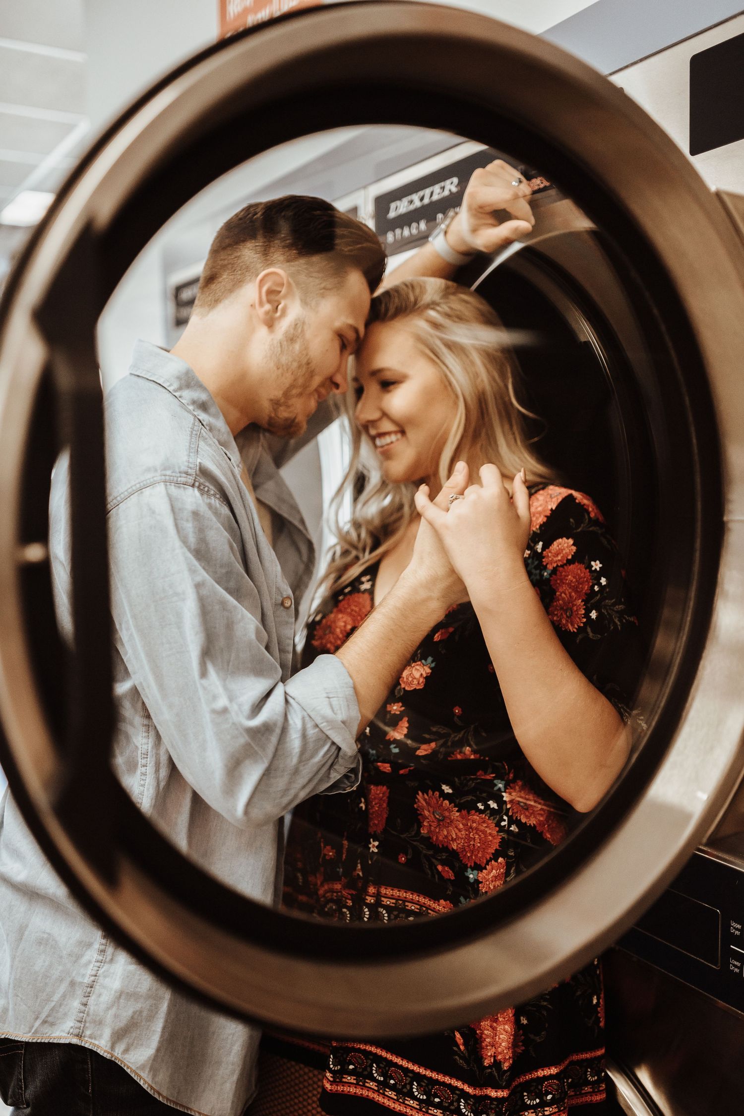 Romantic couple sharing an intimate moment viewed through the frame of a round dryer door at a laundromat.