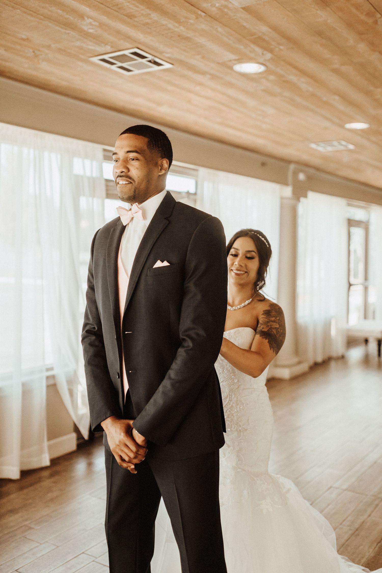 A bride and groom share a tender first look moment in a sunlit venue with wooden floors and sheer white curtains.