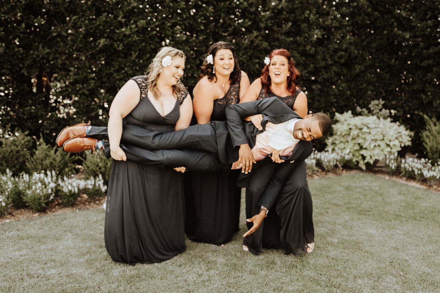 Four people in coordinating black and gray dresses pose playfully together on a garden lawn.