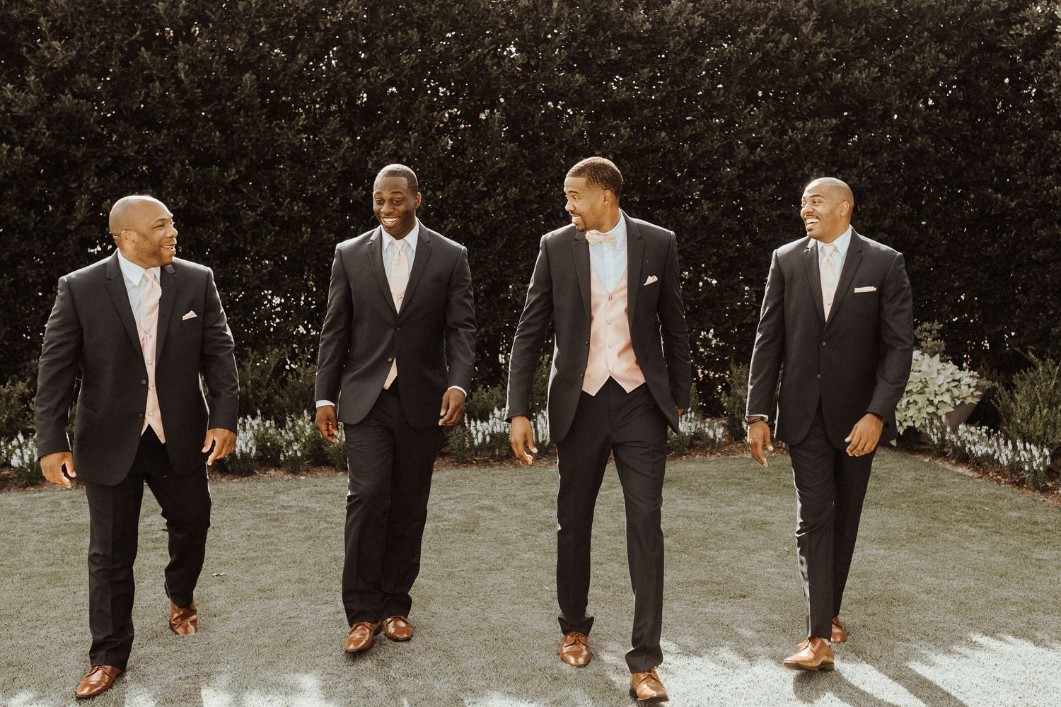 Four groomsmen in stylish black suits walk together on a path in front of a garden hedge on a sunny day.