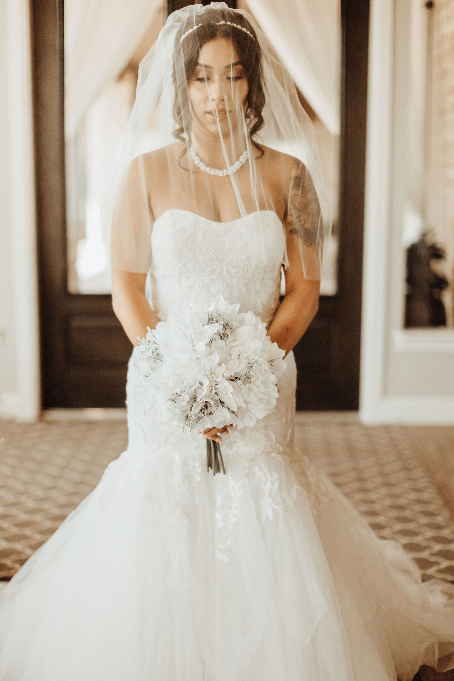 Bride in strapless mermaid wedding gown with tulle veil holds white floral bouquet in sepia-toned portrait.