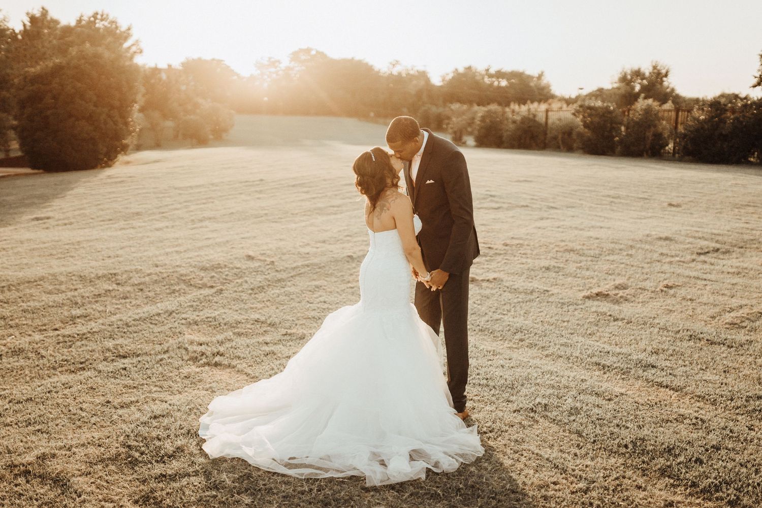 A couple in wedding attire stand close together in a sunlit field during golden hour.