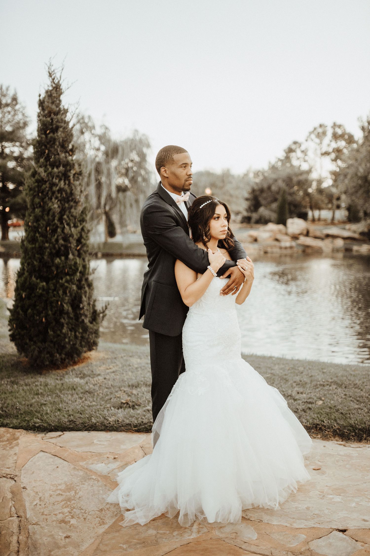 A couple in wedding attire embrace during their golden hour photoshoot by a tranquil lake with cypress trees in the background.