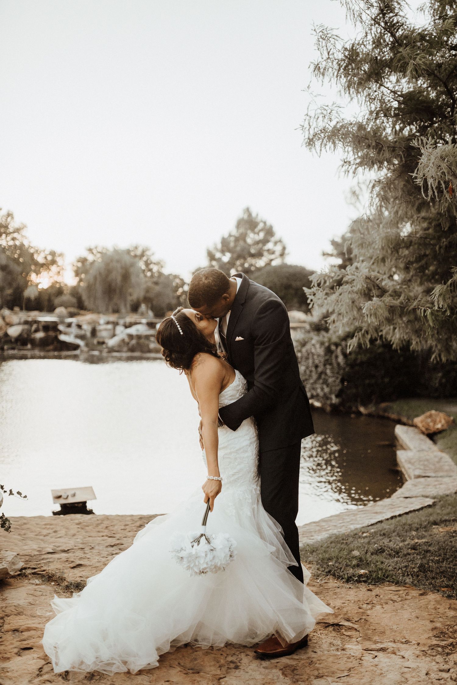 A romantic couple embraces in a dramatic dip kiss by a serene lake at sunset, with the bride wearing a flowing white gown.