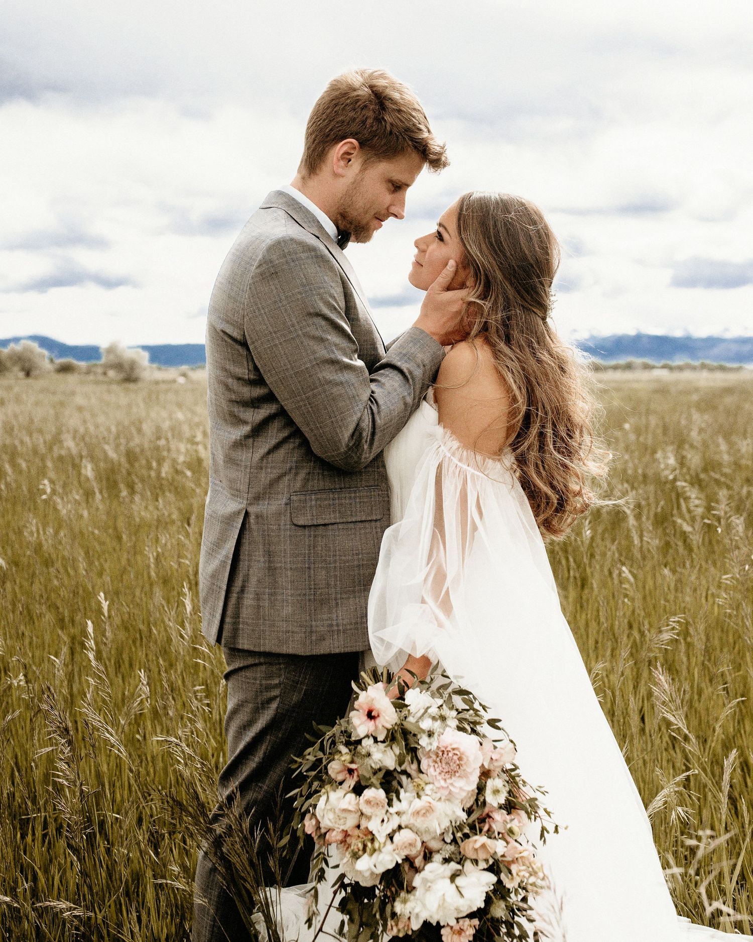 A romantic couple in wedding attire embraces in a rustic wheat field with mountains in the background and a floral bouquet.