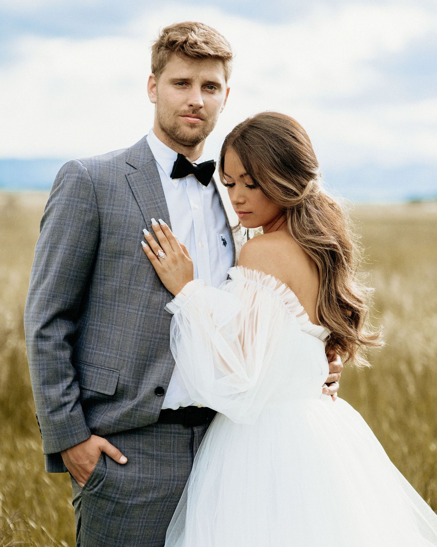 A romantic wedding couple embraces in a golden field under blue skies, the bride wearing a flowing white dress.