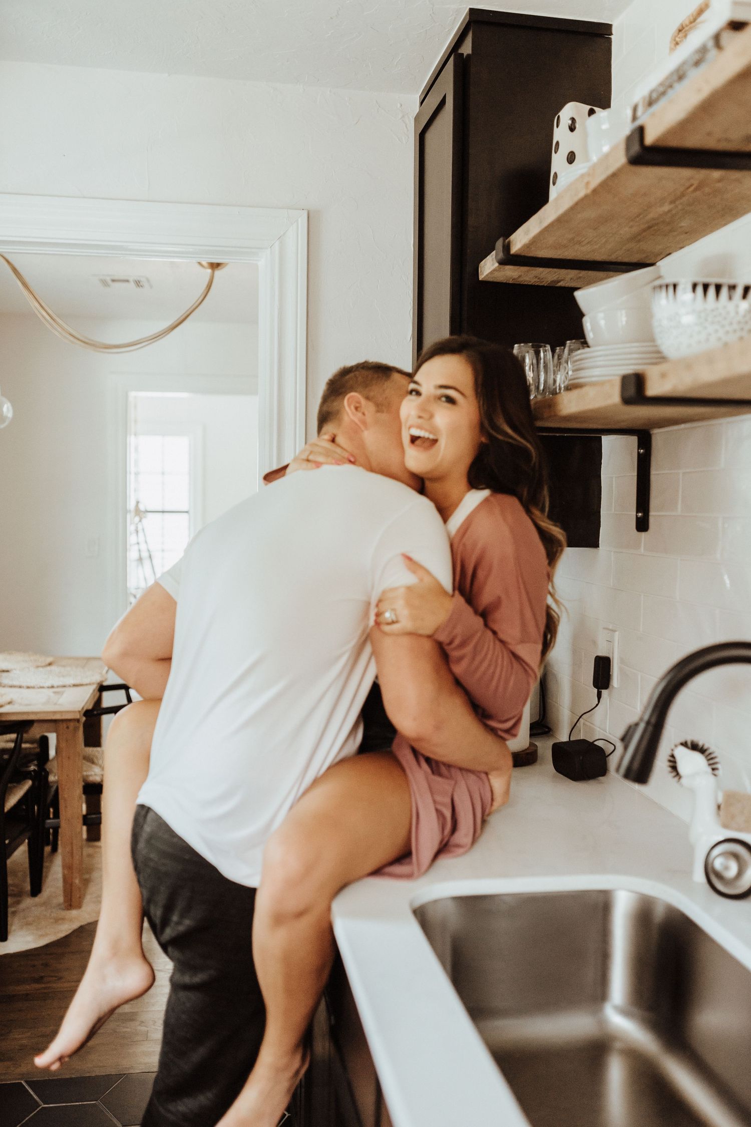 A candid kitchen moment captures a joyful embrace as a couple share a romantic and playful hug by the sink counter.