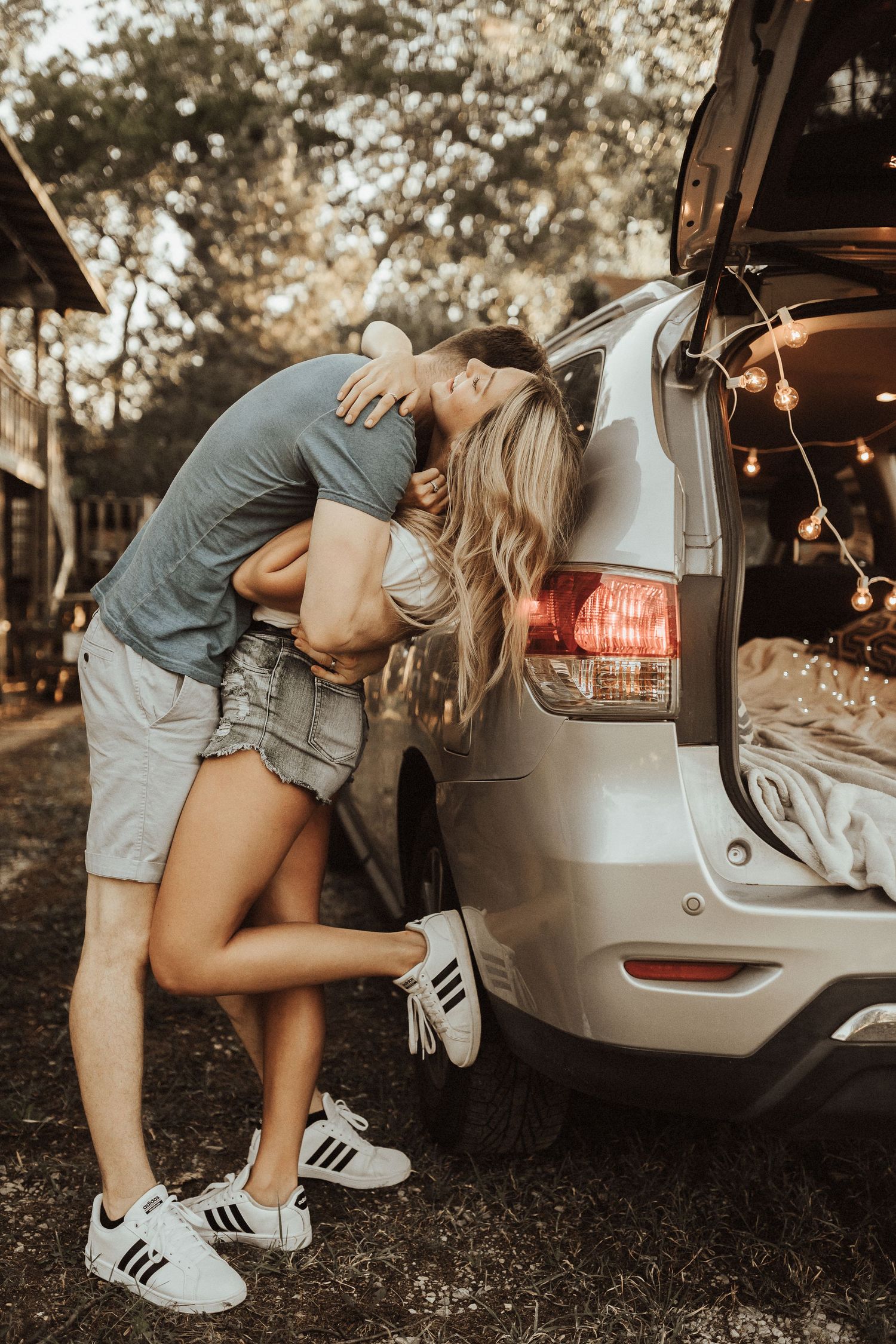 Couple embraces near the trunk of a silver SUV in a romantic outdoor photoshoot with warm lighting.