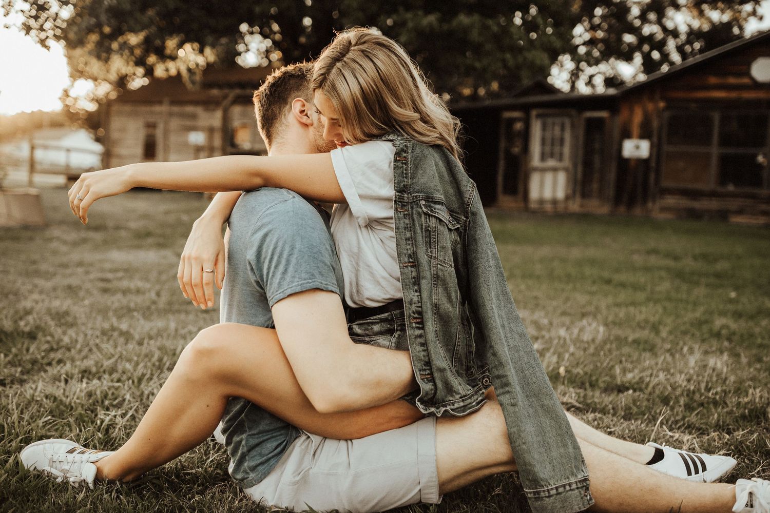 Intimate outdoor embrace between two people sitting on grass at sunset with rustic buildings in background.