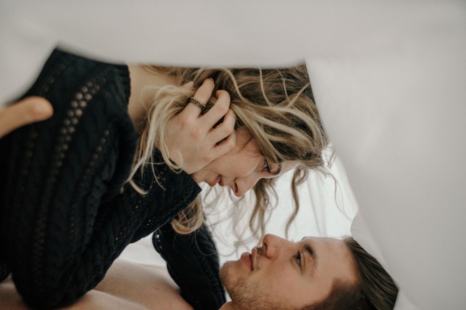 Intimate couple lying down together with woman in black cardigan leaning over partner in tender moment.