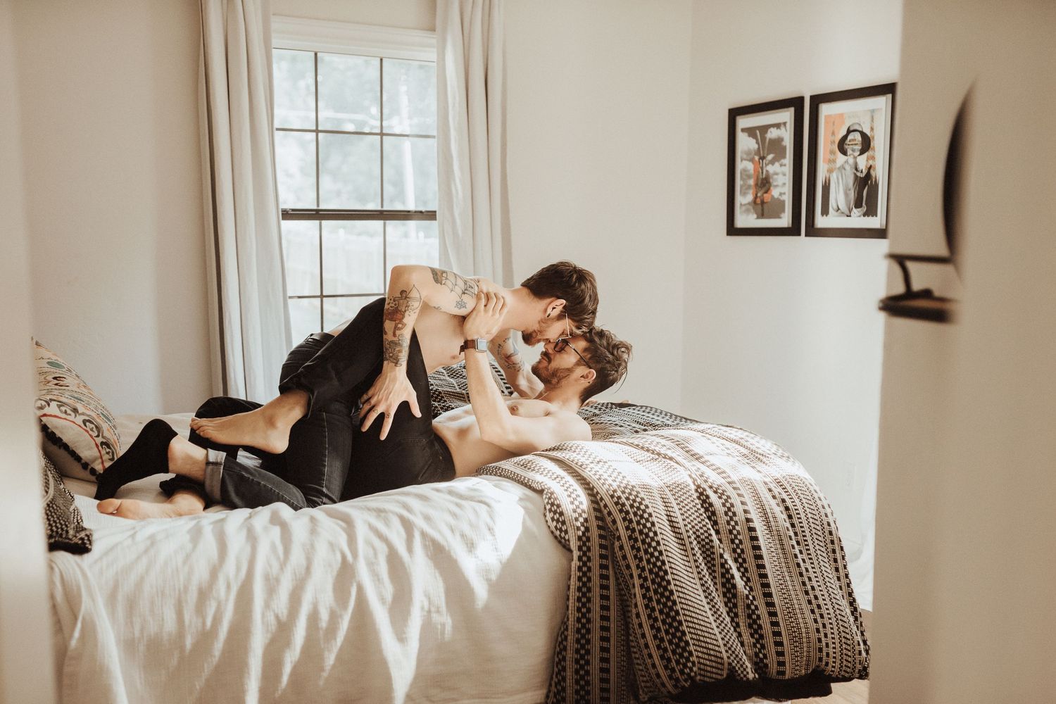 A couple embraces on a bed with white sheets and a knit blanket in a sunlit bedroom with framed photos on the wall.