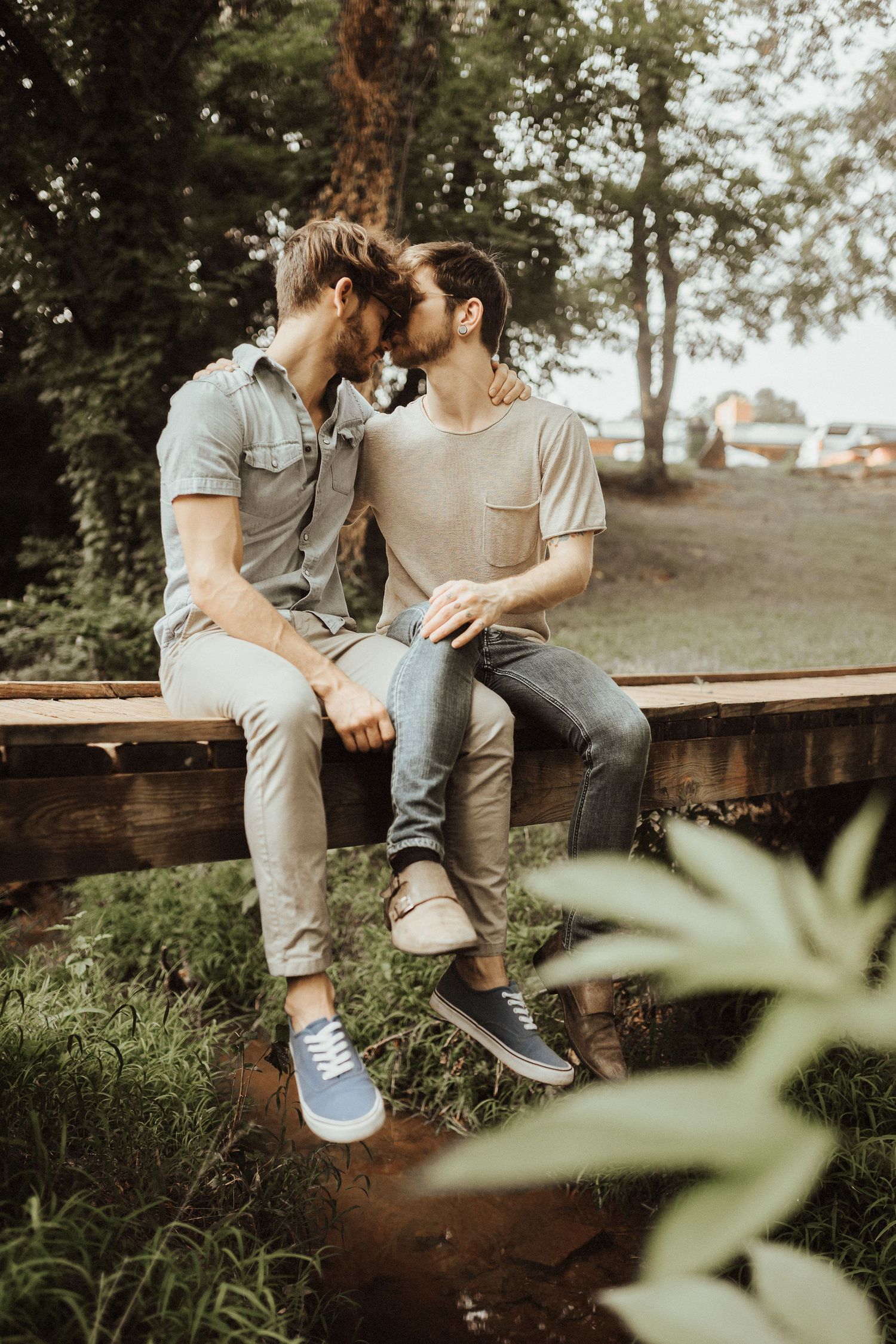 A couple in casual attire share an intimate moment while sitting together on a wooden bench in a natural outdoor setting.