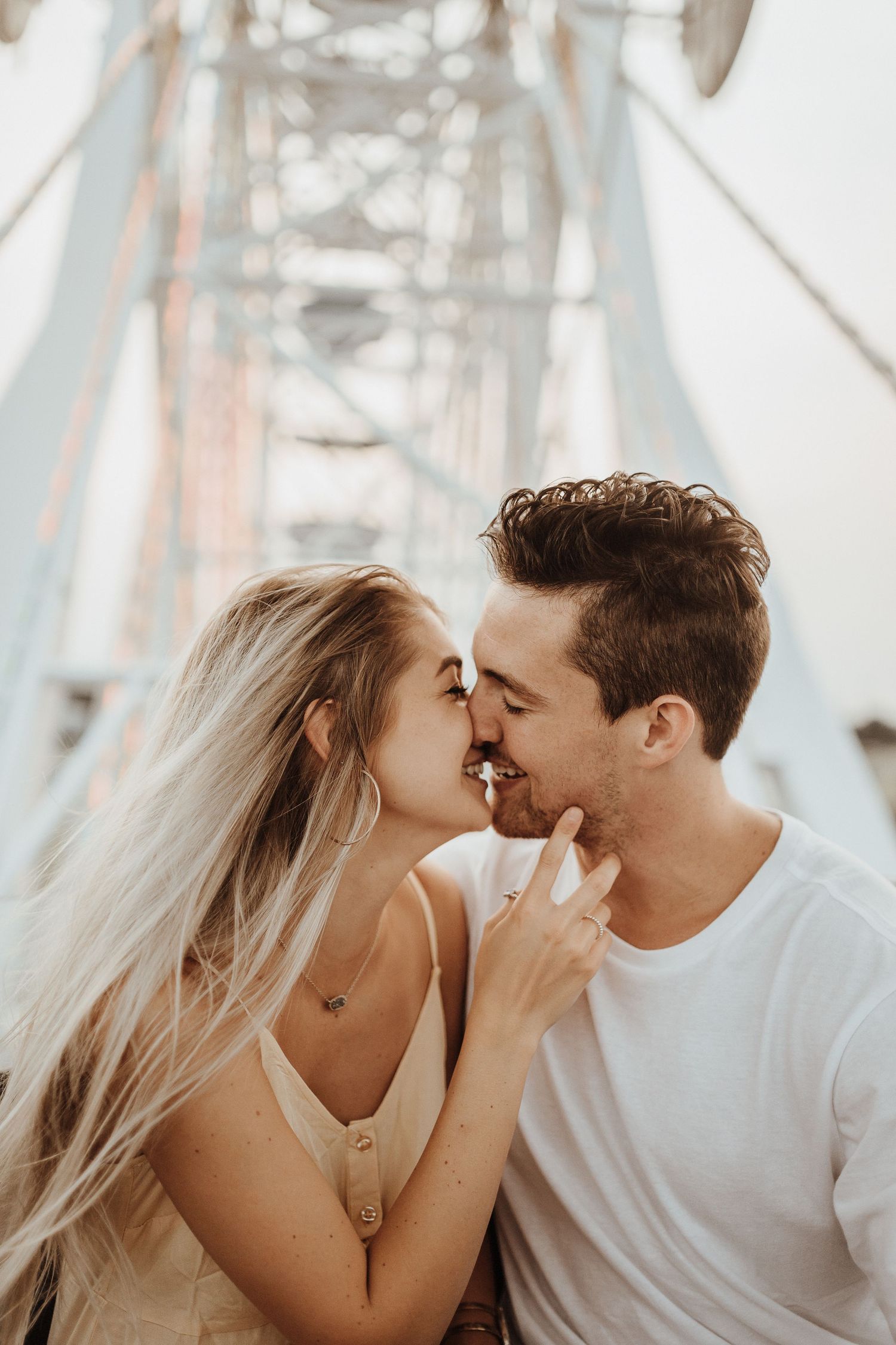 A loving couple shares an intimate moment kissing against a sunlit bridge backdrop.