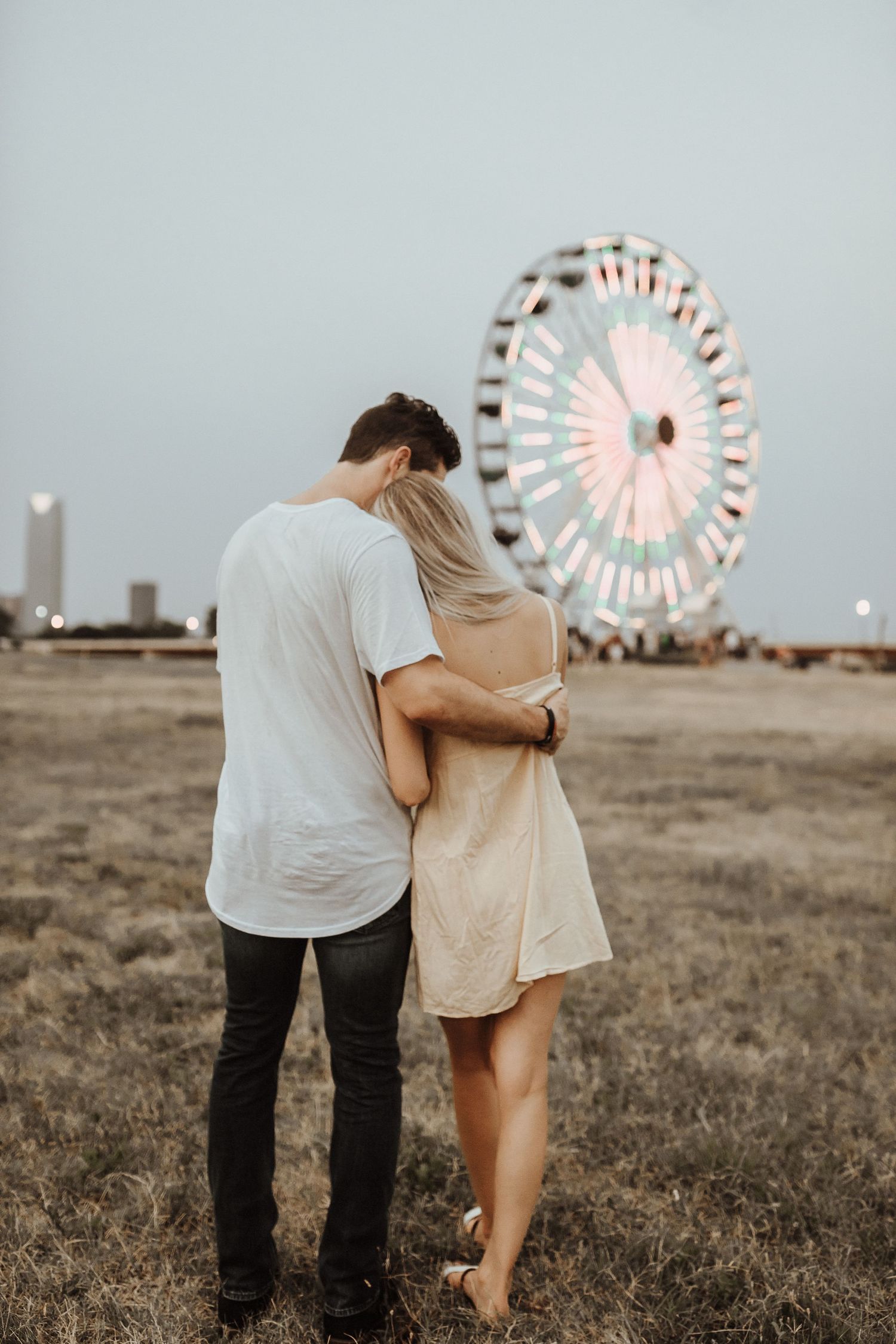 Romantic couple embraces at sunset with illuminated ferris wheel in background on grassy field.