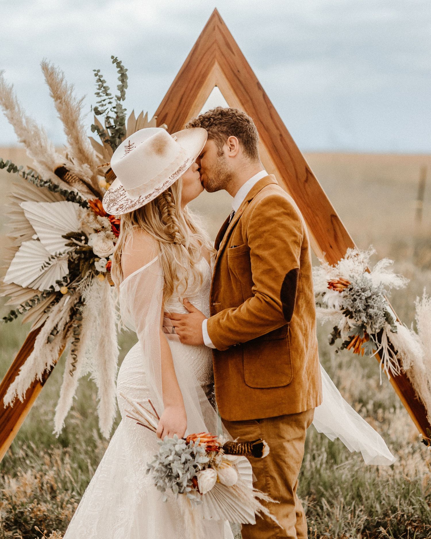 Rustic boho wedding ceremony features wooden triangle arch decorated with pampas grass and dried flowers in warm earth tones.