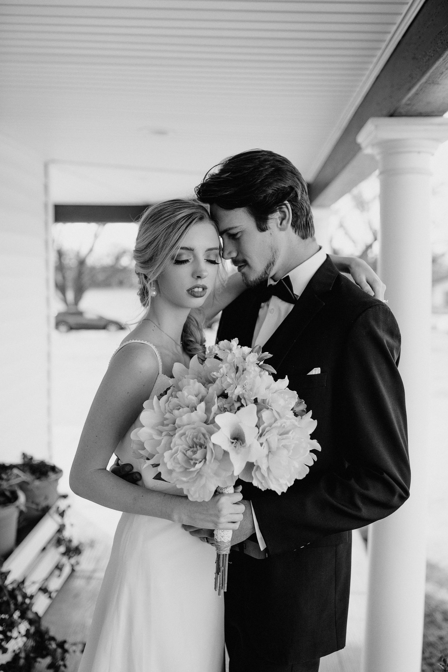 Classic black and white wedding photo showing couple kissing on a porch with a beautiful floral bouquet.