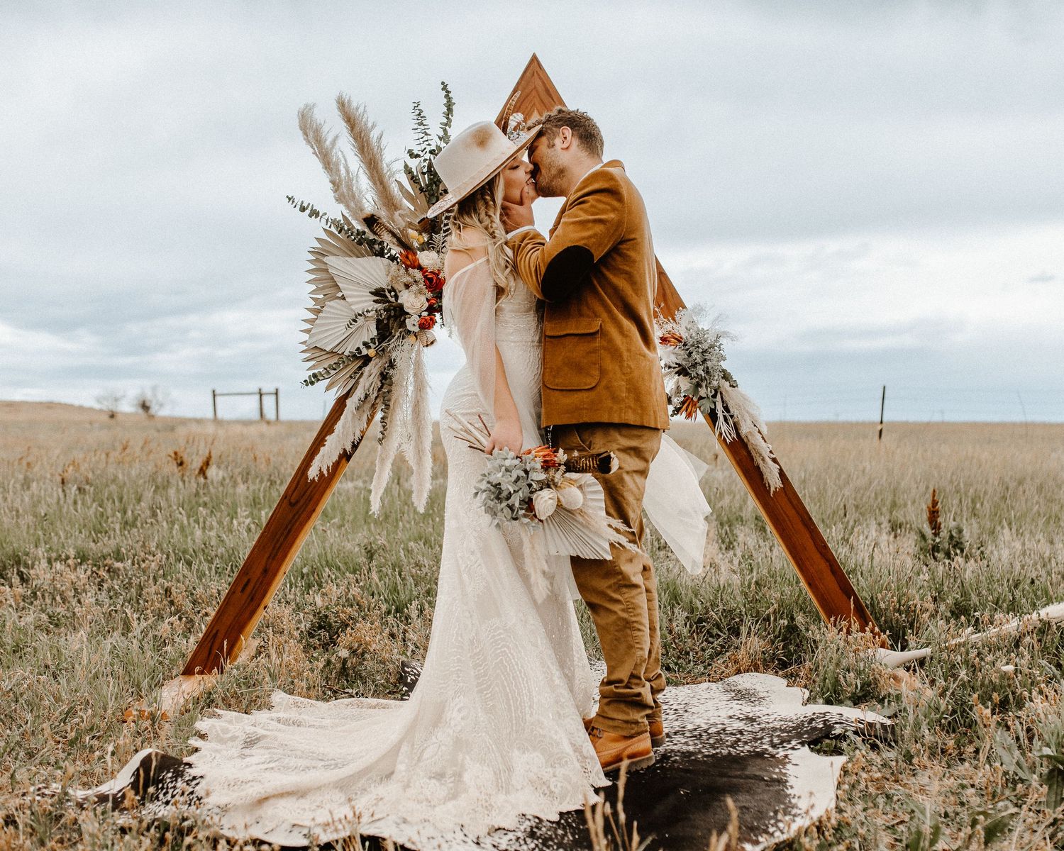 Rustic outdoor wedding ceremony with bohemian wooden triangle arch decorated with pampas grass and dried flowers in prairie field.