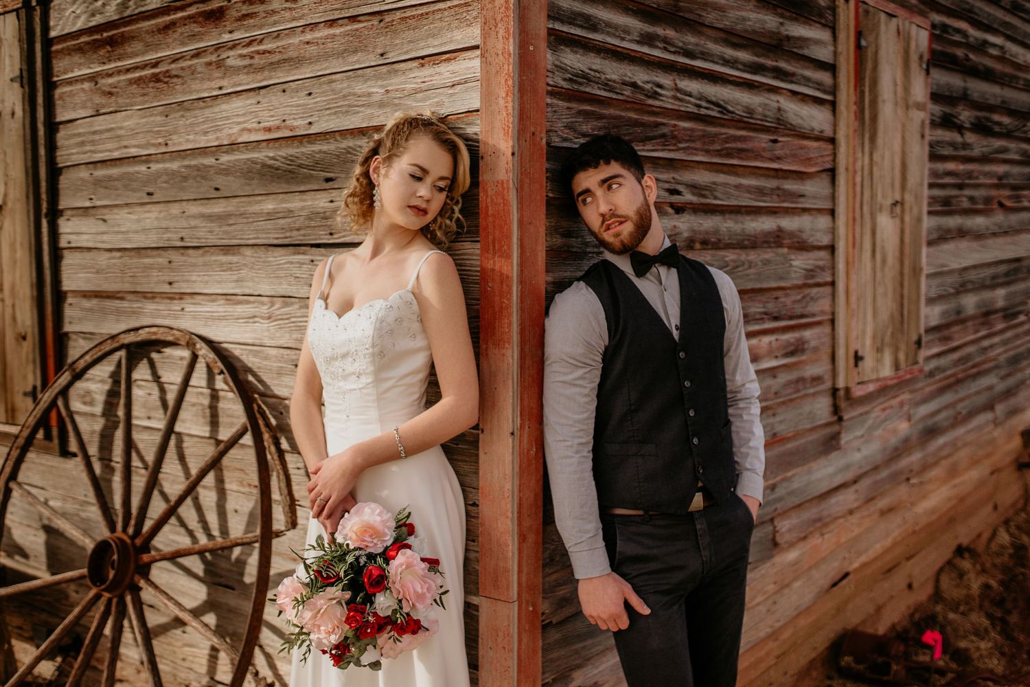 Rustic barn wedding portrait with couple in formal attire posing by wooden wall and vintage wagon wheel.