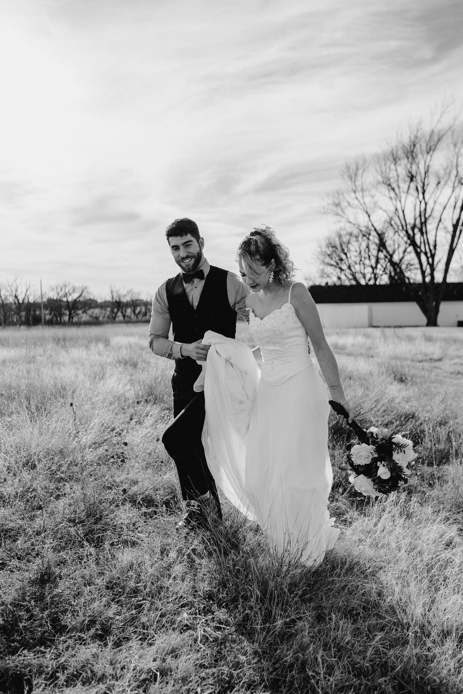 A black and white wedding photo shows couple walking together in a field while holding a bouquet.