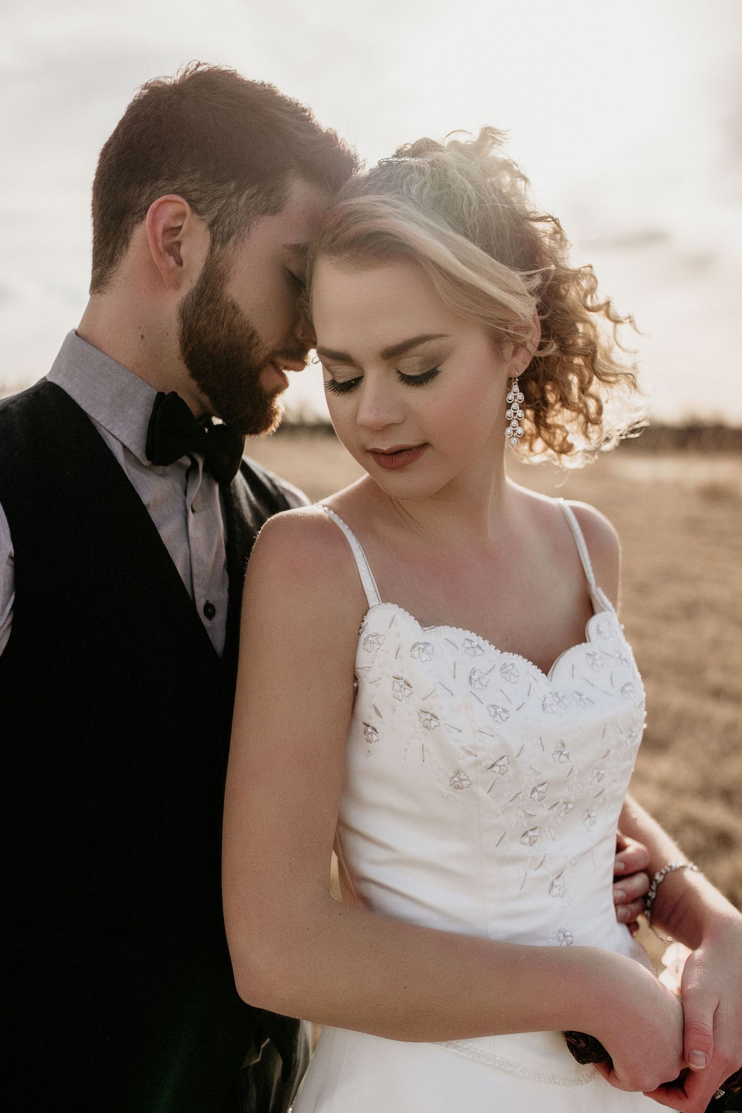 A romantic wedding photo of a couple in formal attire standing close together at sunset with soft lighting.