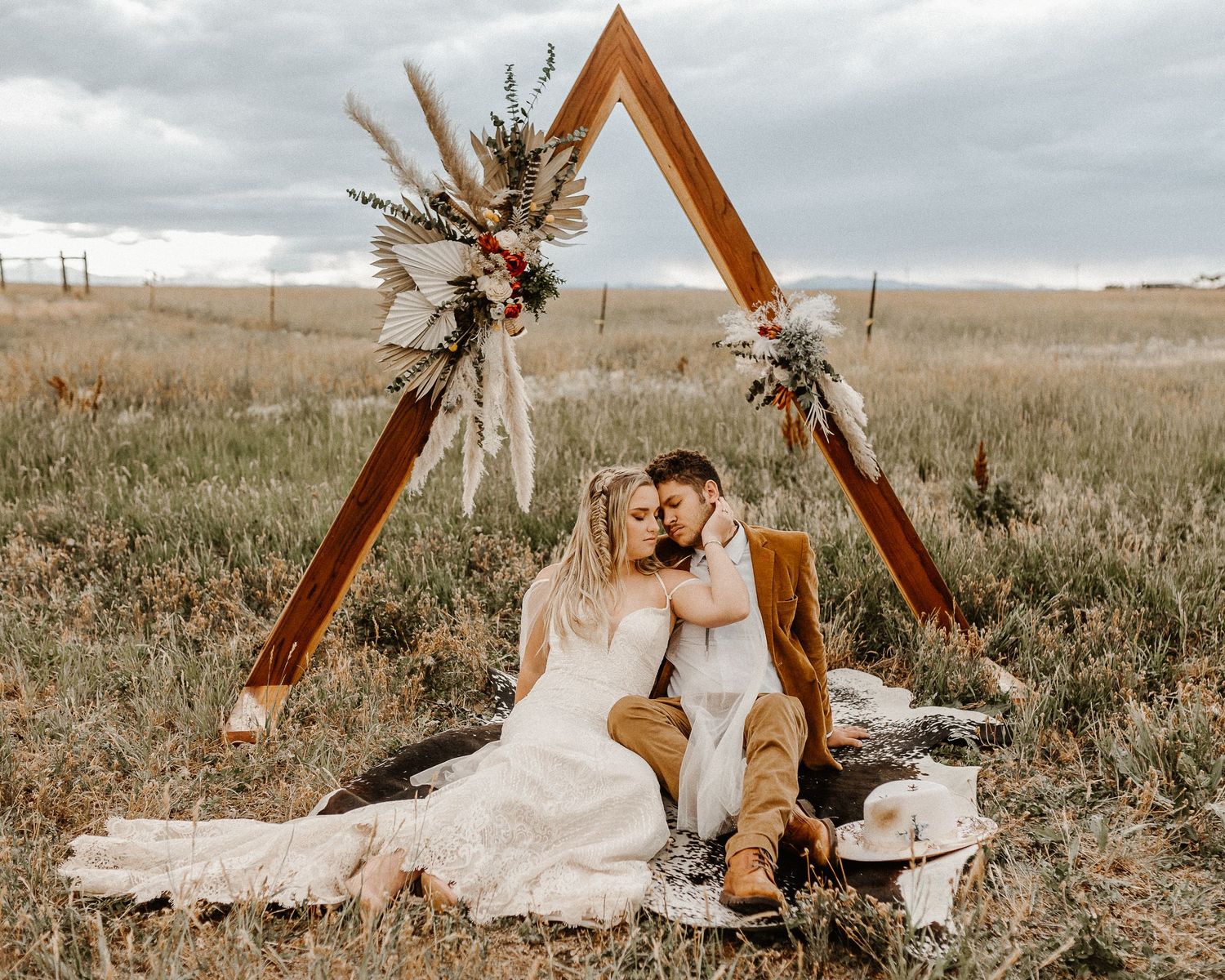 Rustic wooden triangle arch with dried floral arrangements stands in golden field under moody cloudy sky.