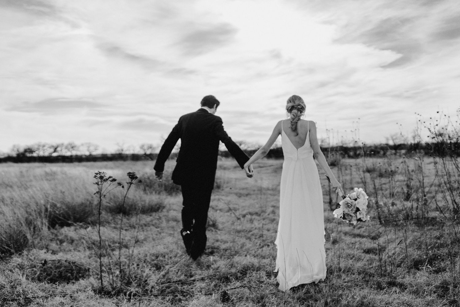 Black and white photograph of a couple in wedding attire walking hand in hand through a scenic field.