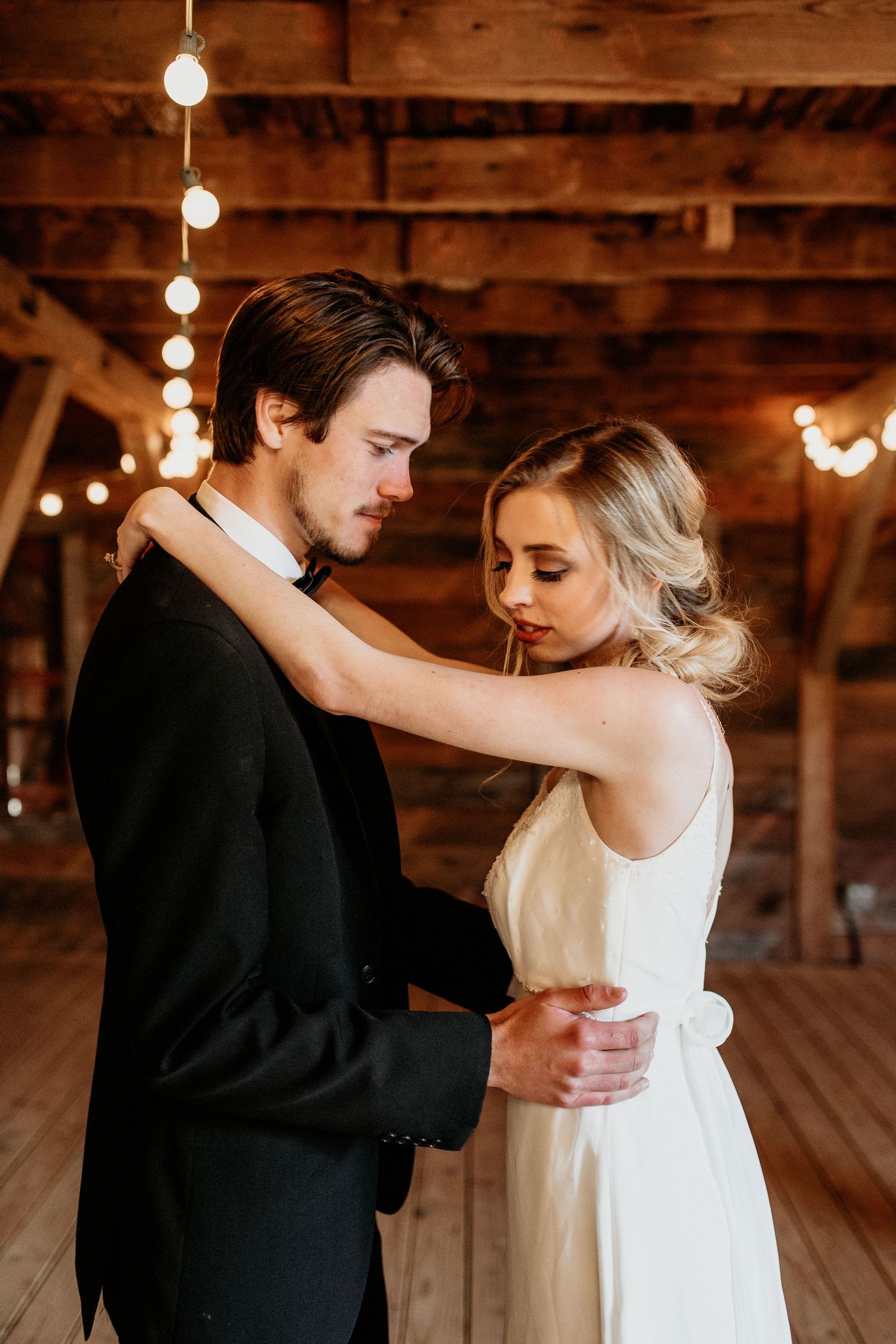A romantic moment between newlyweds dancing in a rustic barn venue with string lights glowing overhead.