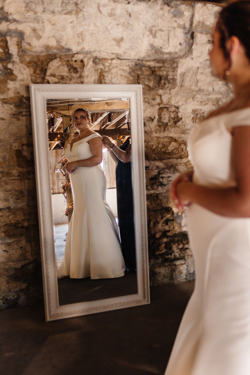 An elegant white wedding dress is reflected in a vintage mirror against a rustic stone wall.