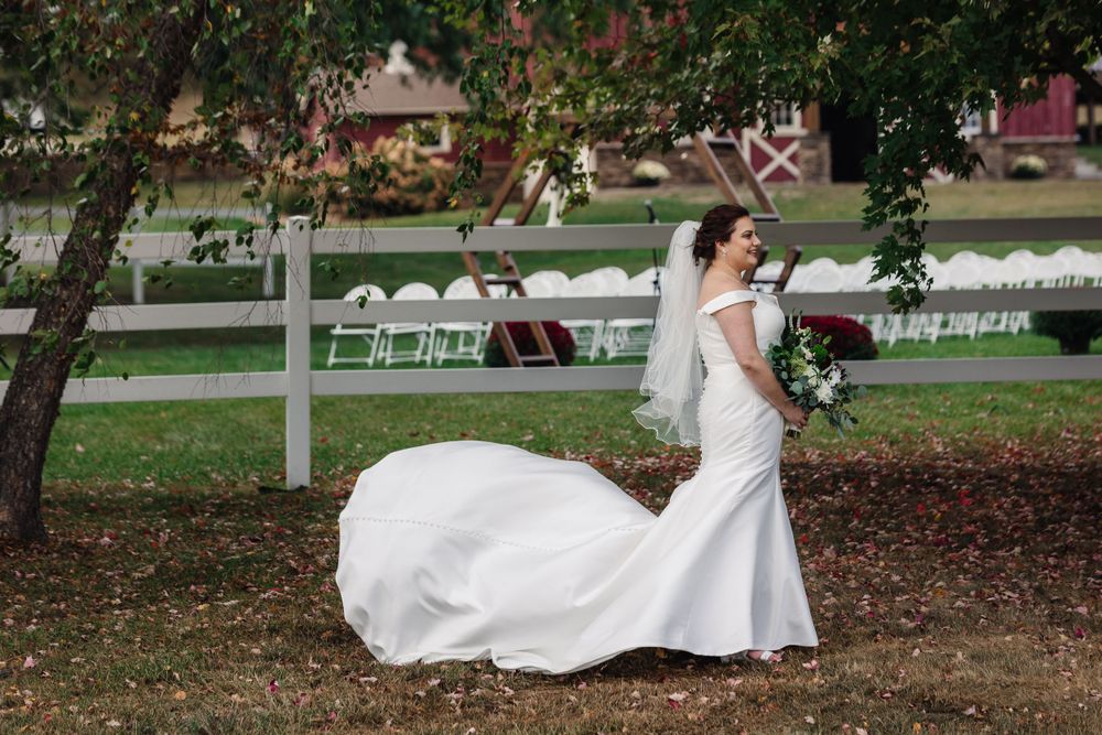 A bride in a flowing white mermaid gown poses outdoors near autumn trees and white chairs.