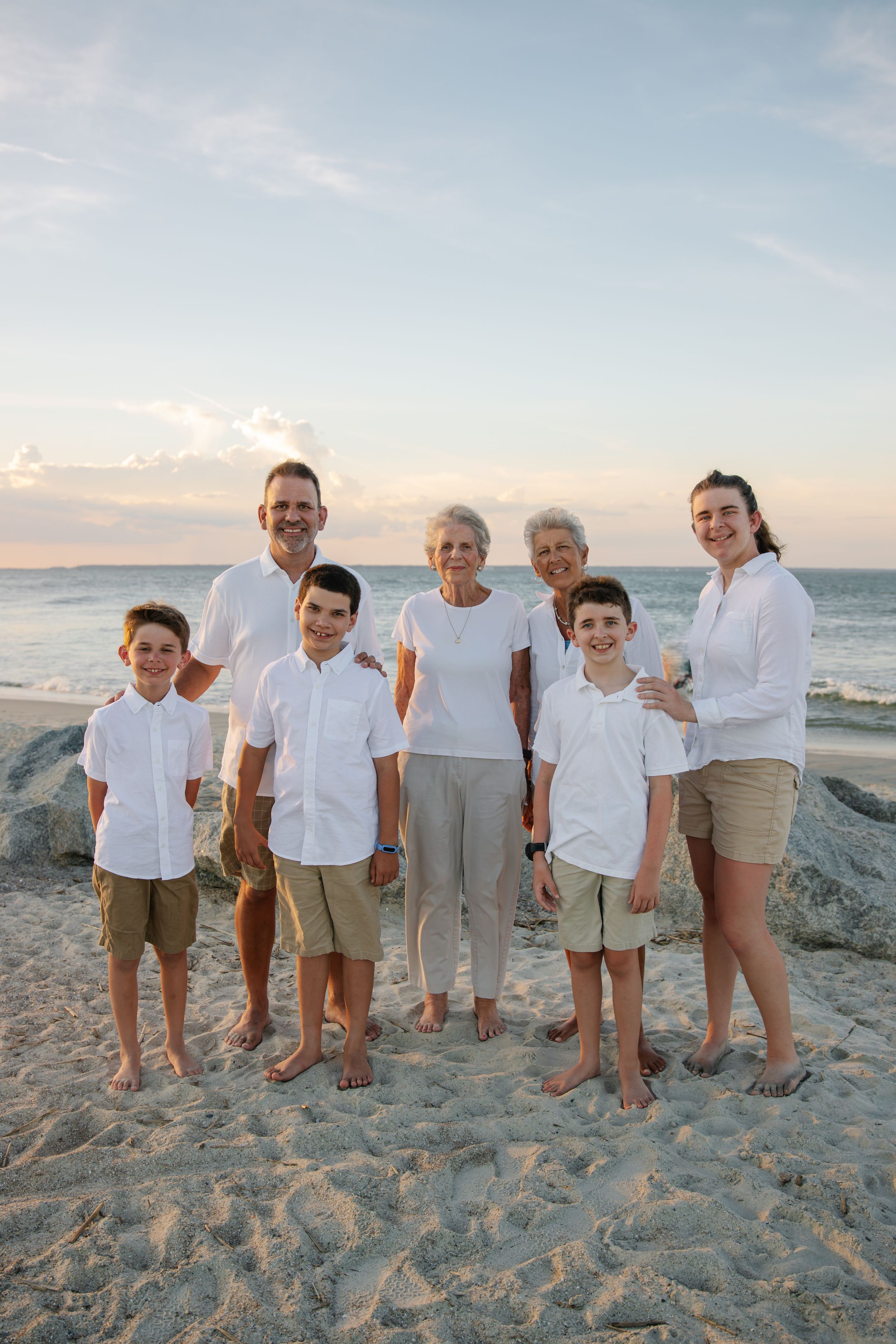 Multi-generational family wearing coordinated white and khaki outfits poses on beach at sunset.