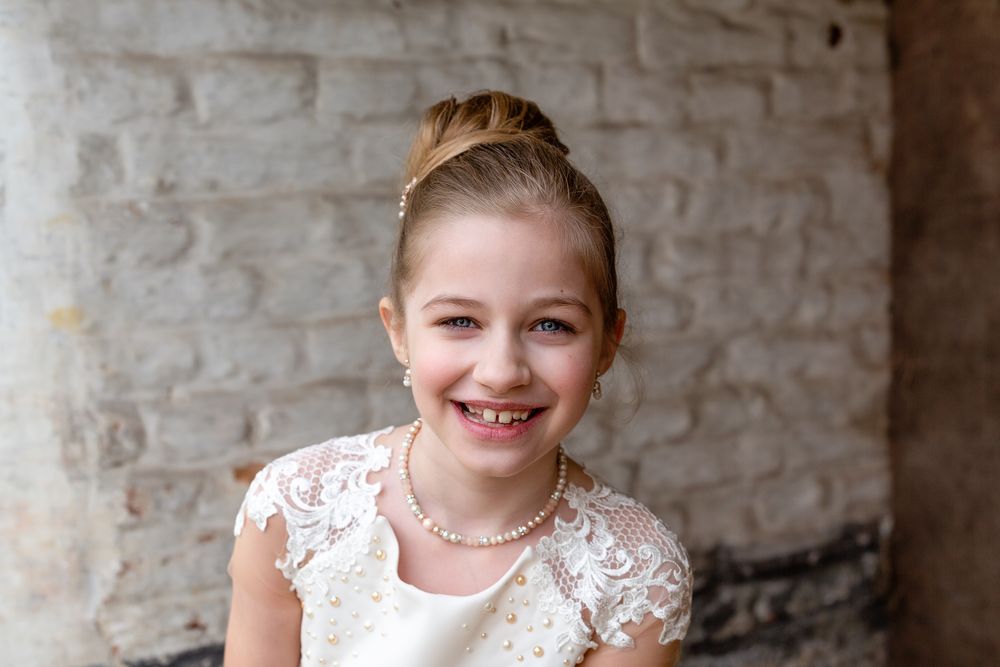 Young person in white lace dress with pearl necklace poses against brick wall.