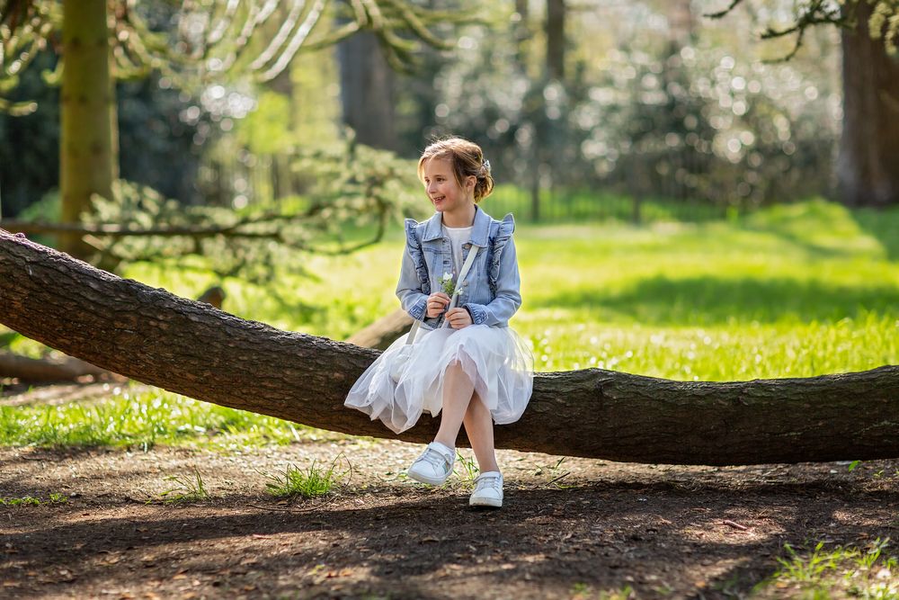 Person wearing denim jacket and white dress sits on fallen tree trunk in sunny garden.
