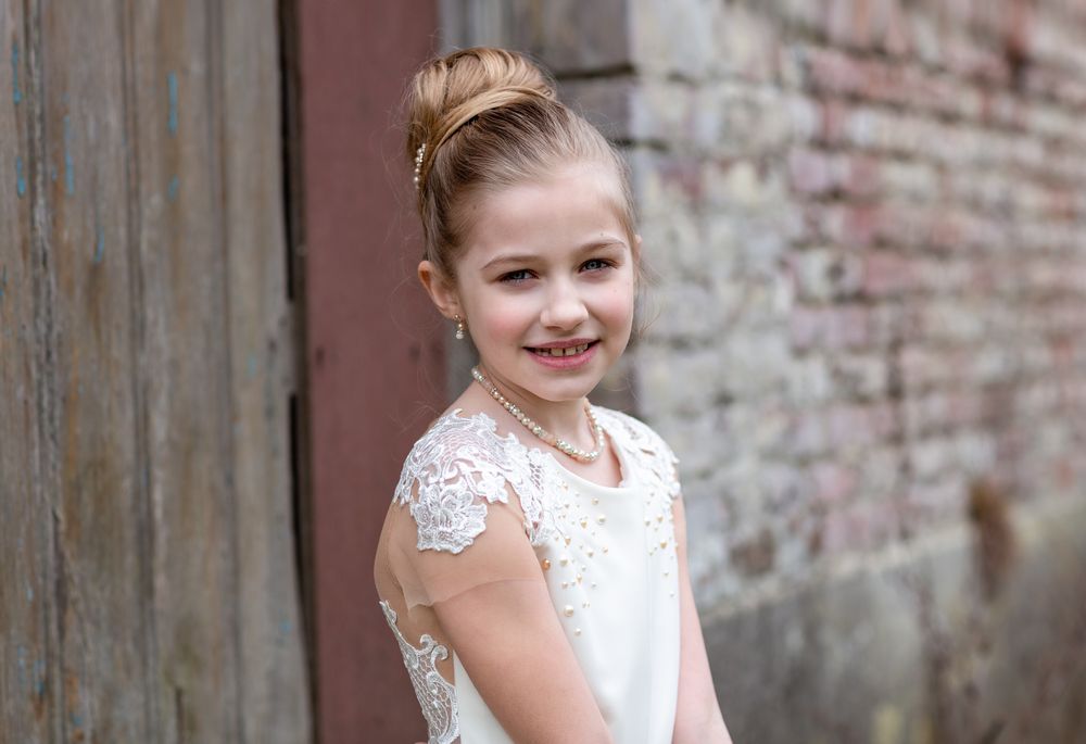 Young person in elegant white lace dress poses against rustic brick wall background.