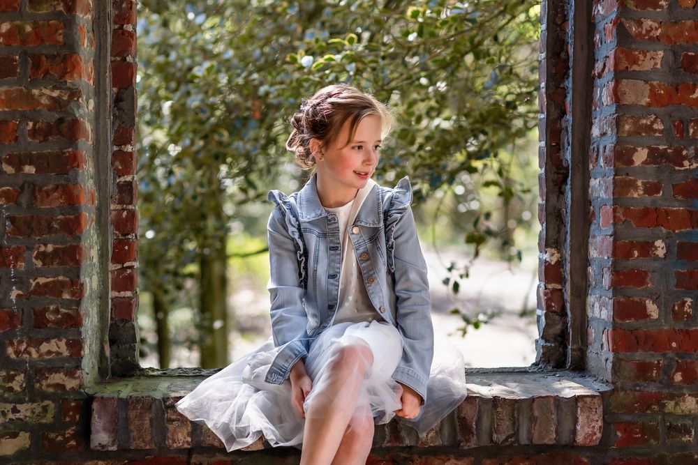 Person in denim jacket and white dress sits on brick windowsill in outdoor garden setting.