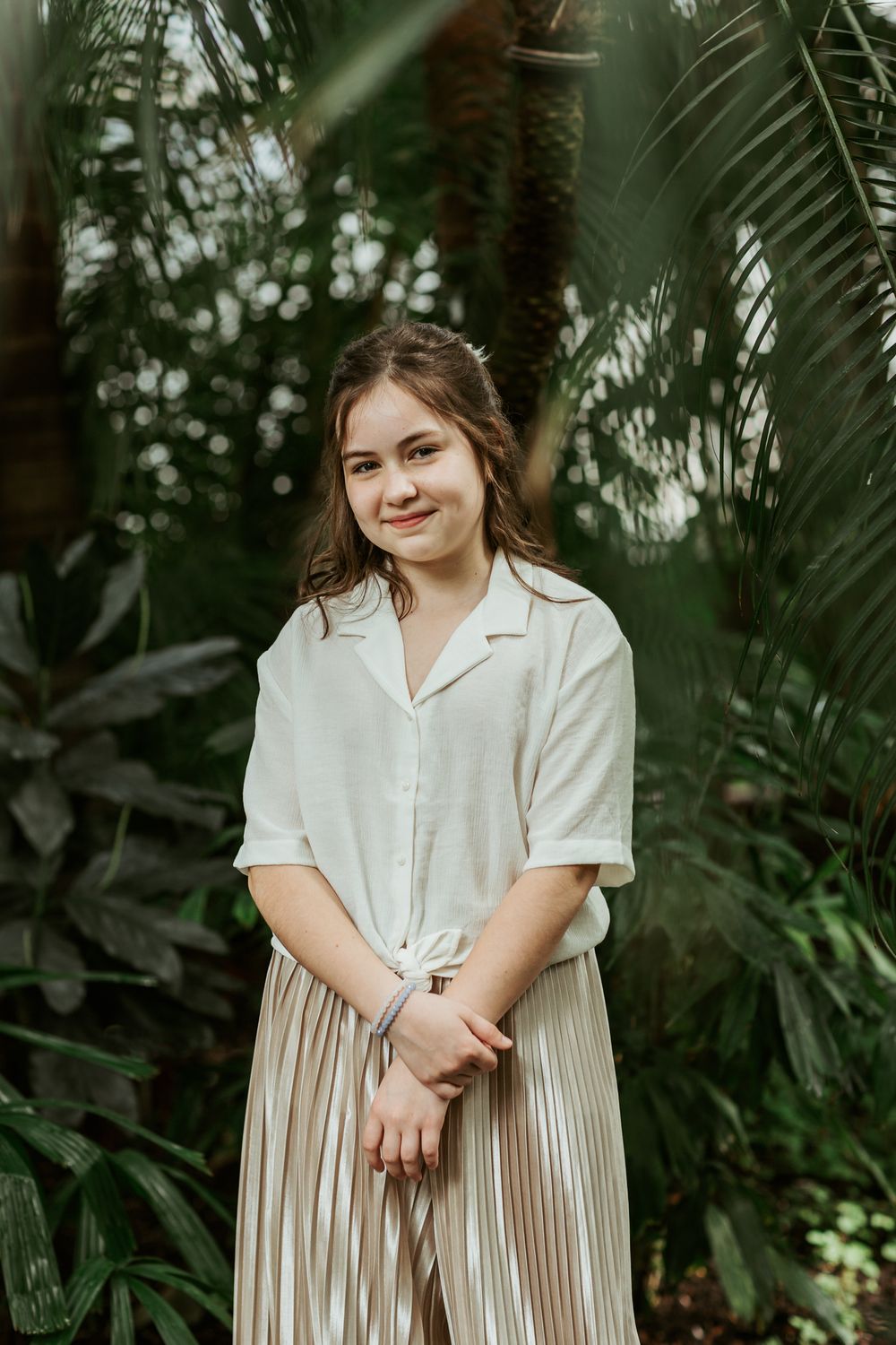 Person in white button-down shirt and pleated skirt poses in lush tropical garden.