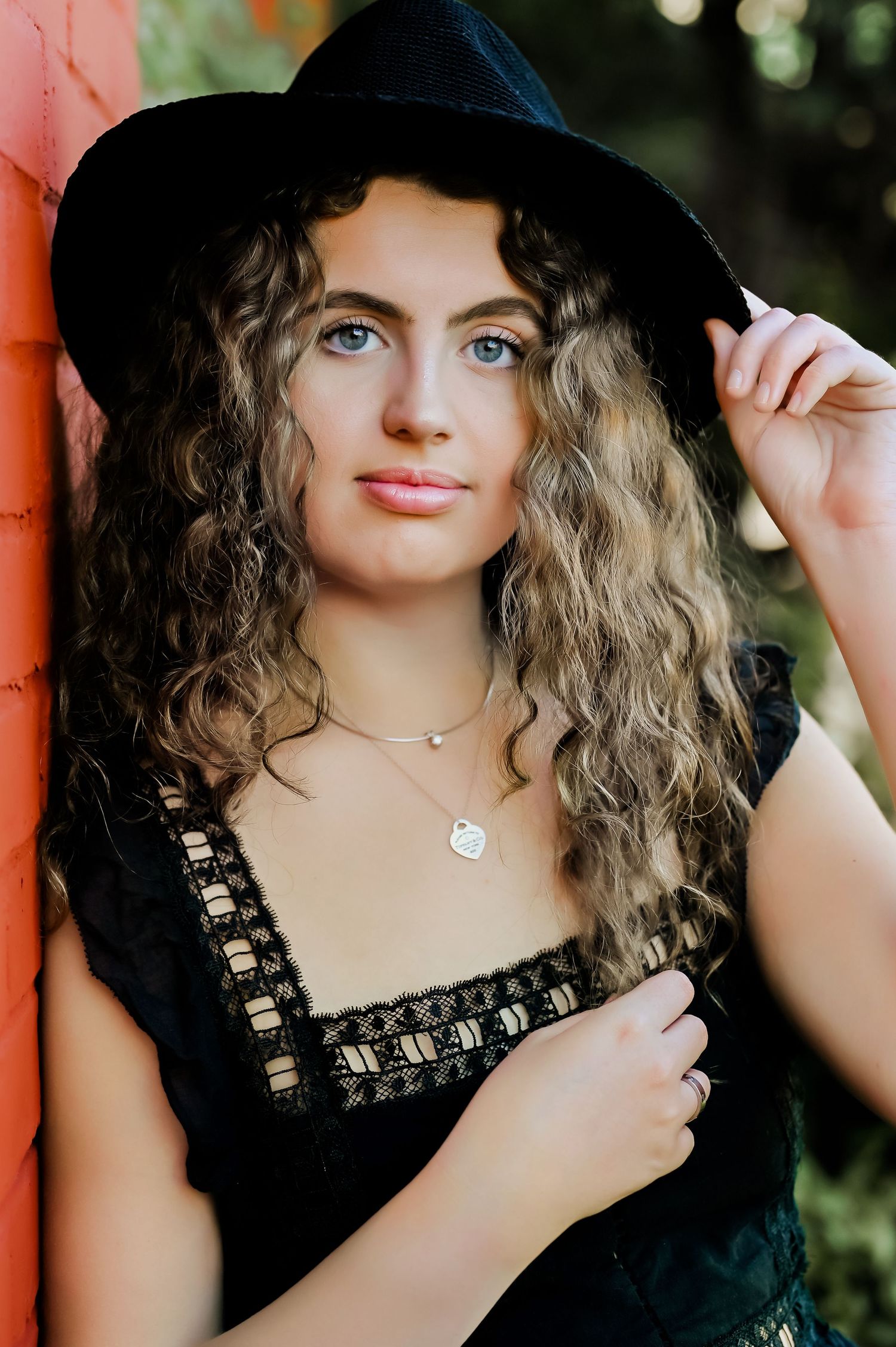 A fashion portrait featuring a black hat and lace top against a brick wall.