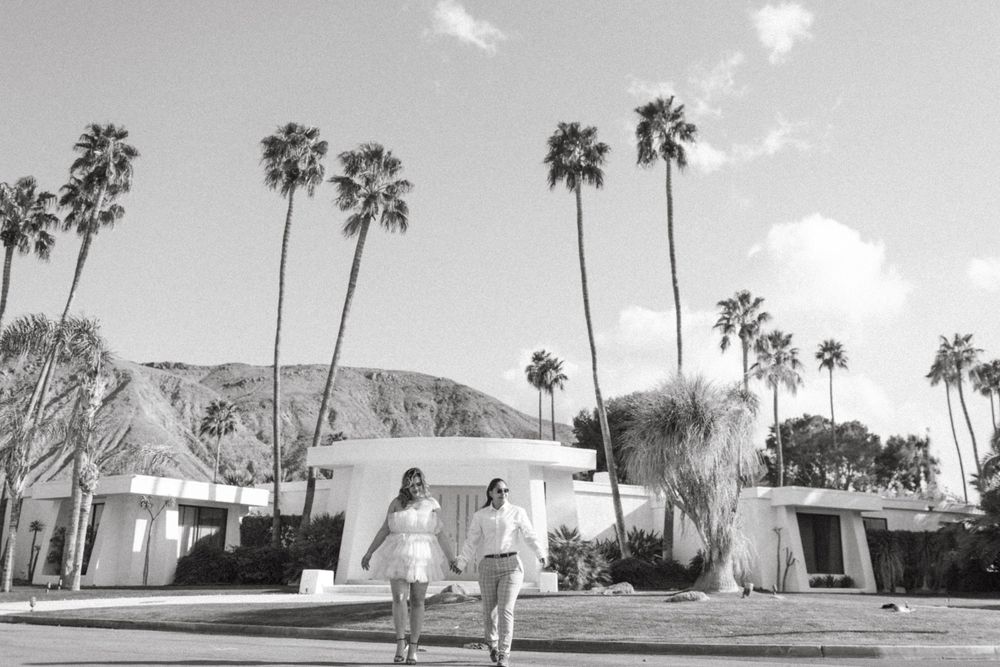 Two engaged women walk holding hands, across the street in front of a Mid-Century Modern Home in Palm Springs