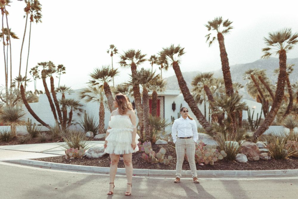 in an urban setting, with palm trees and a Mid-Century Modern home in the background, Two women look off into distance