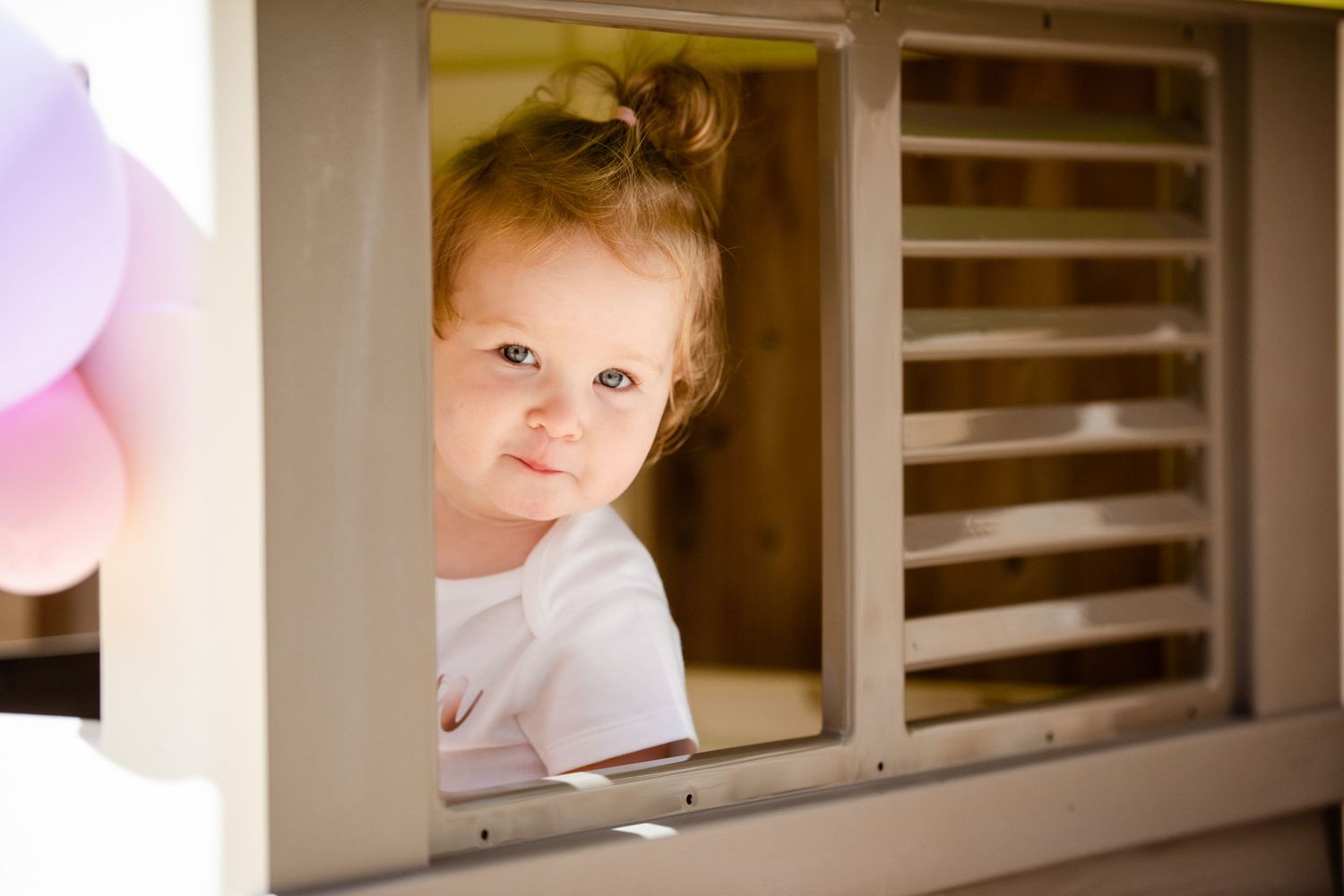 Small child peeks through window with pink balloons in frame.