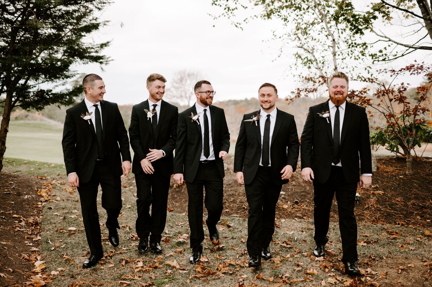 Groomsmen in black suits walk together on autumn path sharing candid moment.