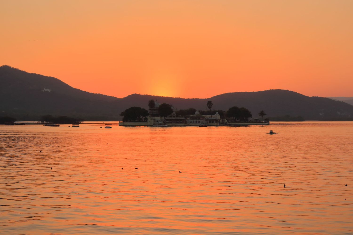Orange sunset over calm lake waters with silhouetted boats and mountains in background.