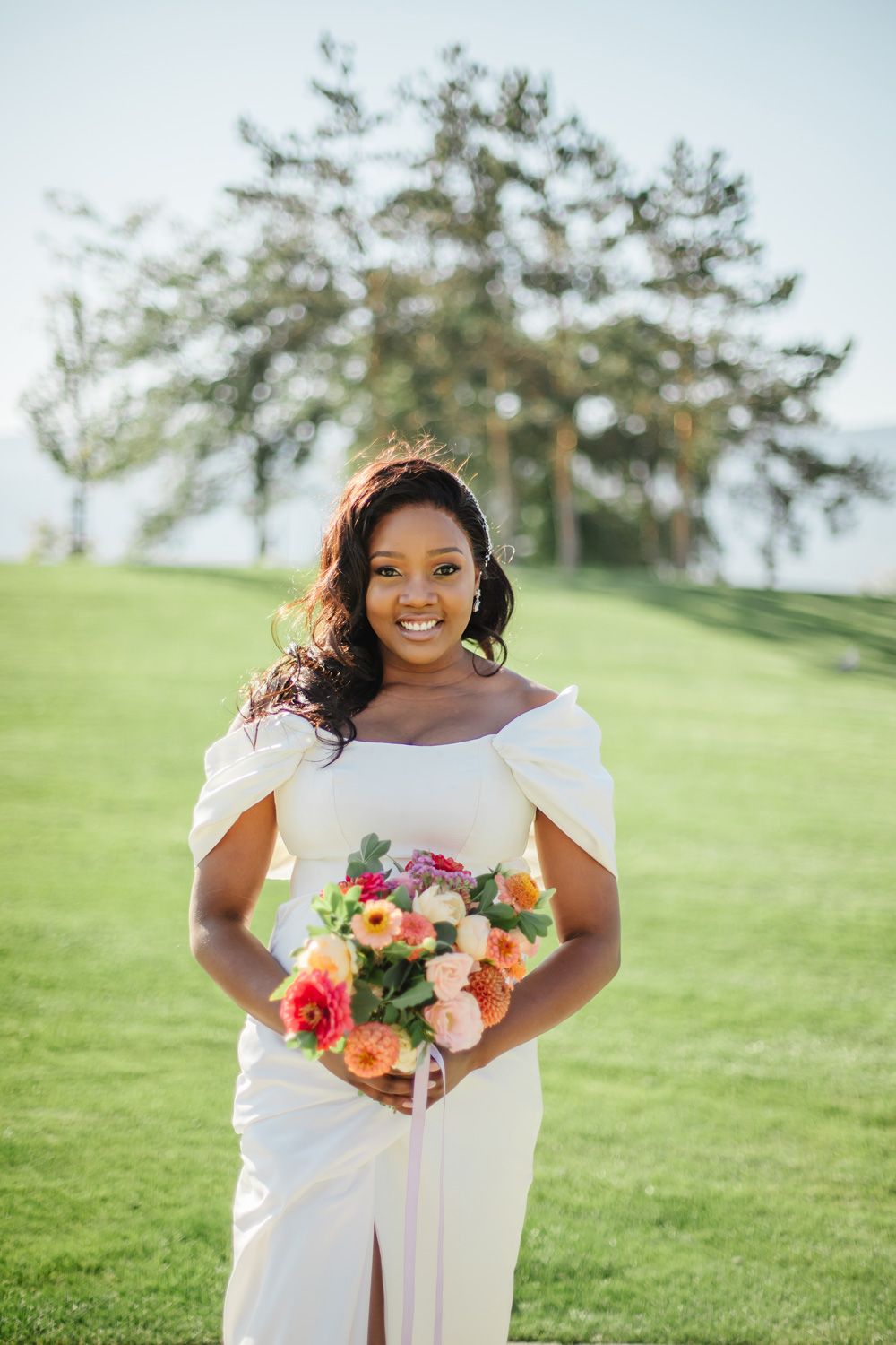 A radiant bride holds a vibrant bouquet of orange and pink flowers on a grassy hillside.