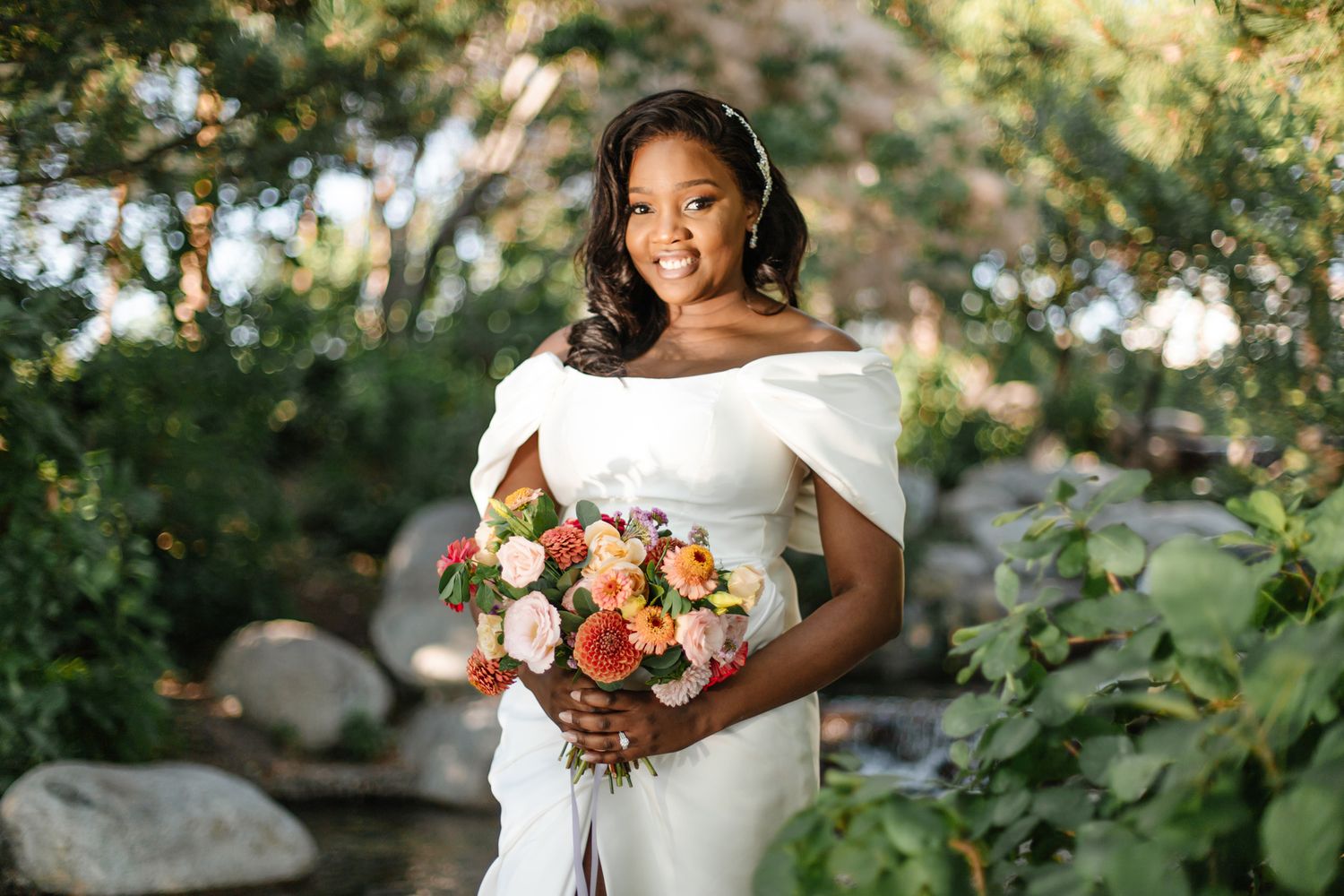 A bride in a white dress holds a vibrant coral and peach bouquet against a lush garden backdrop.