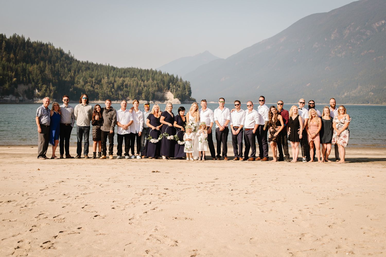 Group photo of wedding party lined up on beach with mountain landscape.