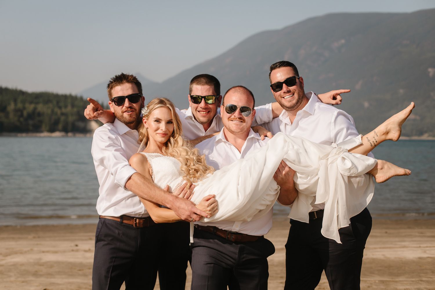 Wedding party poses playfully on a scenic beach with mountains in the background.