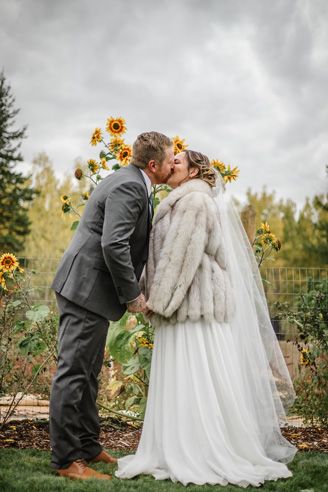 A wedding couple shares a romantic kiss in a garden with blooming sunflowers and autumn foliage in the background.
