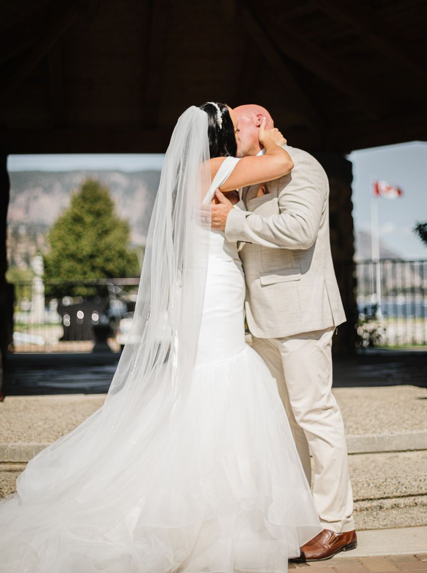 A romantic wedding kiss under an archway with a flowing white veil and beige suit.